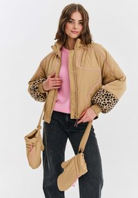 Žena v béžovej prešívanej bunde s leopardími rukávmi, ružovom svetri, čiernych džínsach a béžových rukaviciach zavesených na rukávoch bundy.