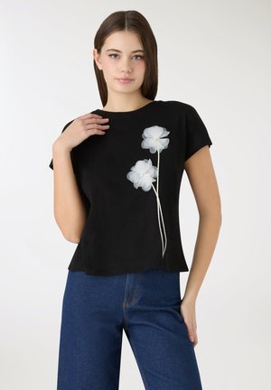 Czarny, krótki top o luźnym kroju, zdobiony dwoma białymi aplikacjami kwiatowymi i teksturowanym wykończeniem. Połączony z ciemnoniebieskimi, szerokimi spodniami jeansowymi.