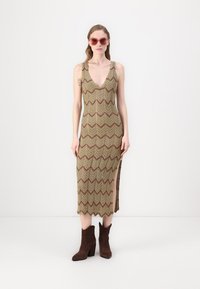 Abito maxi in maglia con motivo a zigzag in tonalità terra, caratterizzato da un profondo scollo a V, spacco laterale e abbinato a stivali alla caviglia marroni.