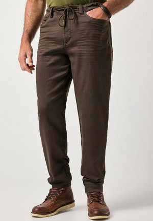 Mężczyzna w brązowych spodniach z sznurkiem i brązowych skórzanych butach, z jedną ręką w kieszeni i częściowo widocznym zielonym topem.