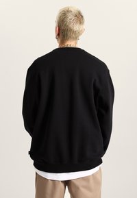Black crewneck sweatshirt made of soft fabric, featuring a relaxed fit and ribbed cuffs, worn over a white shirt and khaki pants.