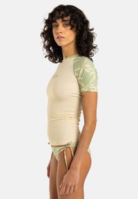 Rash guard de manga corta en color beige claro con un patrón de hojas verdes en las mangas; diseño ajustado, textura suave, combinado con un bikini de lado atado a juego.