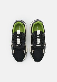 Ett par svarta sneakers med vita och beige detaljer, svarta snörningar, grön innersula och vit tunga, sett ovanifrån på vit bakgrund.