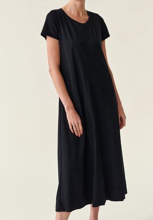 Femme portant une robe midi noire ample à manches courtes, debout devant un fond clair uni.