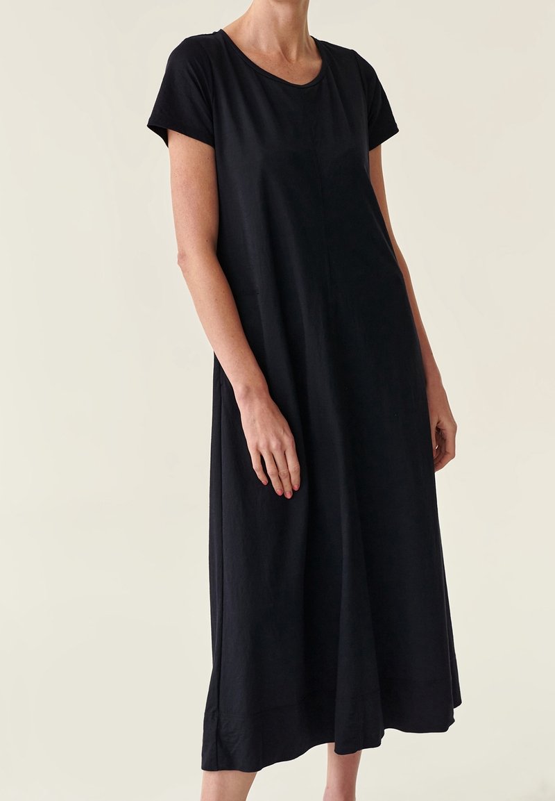 Femme portant une robe midi noire ample à manches courtes, debout devant un fond clair uni.
