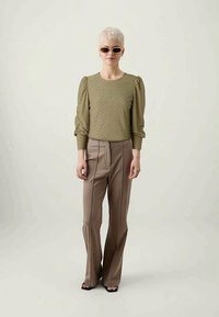Suéter de malha verde-oliva com mangas bufantes, combinado com calças castanhas de cintura alta e corte ajustado. A modelo usa óculos de sol e sandálias pretas.