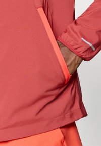 Χέρι που φοράει κόκκινη αθλητική ζακέτα με ανακλαστική ρίγα στο μανίκι, χέρι τοποθετημένο σε μπροστινή τσέπη με φωτεινές κόκκινες λεπτομέρειες.