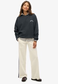 Donkergrijze oversized hoodie met "SUPERDRY" logo, gelaagd over een gestreepte blouse met kraag. Gecombineerd met ivoorkleurige wijde broek en loafers.