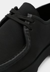 Chaussure noire en matériau synthétique lisse, à bout rond et à couture simple. Présente des lacets plats et une surface texturée.