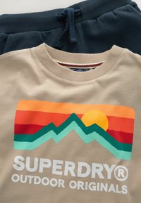 Beige bavlněné tričko s multicolorovým grafickým motivem hor a slunce, se slovy "SUPERDRY OUTDOOR ORIGINALS" v tučných bílých písmech na spodní části.