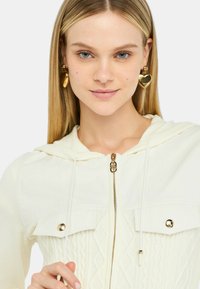 Felpa zip con cappuccio di colore crema, caratterizzata da un design a maglia testurizzata, con dettagli in metallo dorato e cappuccio regolabile; indossata con grandi orecchini a forma di cuore dorati.
