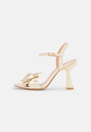 Beige højhælt sandal med blomsterdekoration på tåremmen og justerbar ankelrem med en guldbuckle.