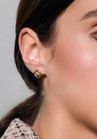 Złoty kolczyk w kształcie obręczy prezentowany na uchu modelki, z gładkim, okrągłym wzorem o wypolerowanym wykończeniu. Subtelny połysk i minimalne zdobienia.