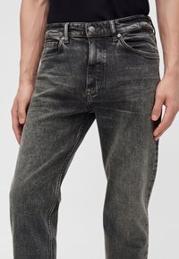 Gros plan d'une personne portant un jean en denim noir délavé et une chemise noire, les mains détendues le long du corps sur un fond clair uni.