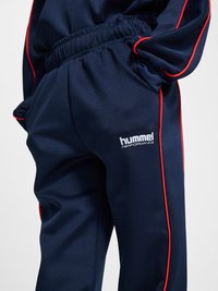 Granatowe spodnie dresowe z czerwonymi lamówkami, noszone z dopasowaną kurtką, z elastycznym pasem i białym logo „hummel performance” na udzie.