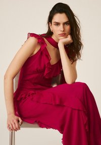 Mujer con cabello largo y oscuro sentada en una silla, vistiendo un vestido sin mangas de color rosa brillante con detalles de volantes, apoyando la barbilla en su mano.