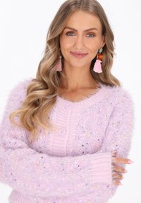 Kobieta z falującymi blond włosami nosi puszysty fioletowy kardigan z guzikami, różowe kolczyki z frędzlami i krzyżuje ramiona, uśmiechając się do kamery.
