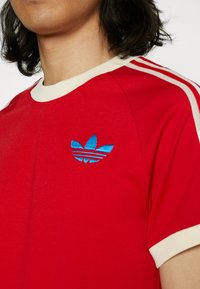 Červené bavlněné tričko s kulatým krémovým výstřihem. Obsahuje modrou vyšívanou značku na levém hrudníku a krémové proužky na ramenou.