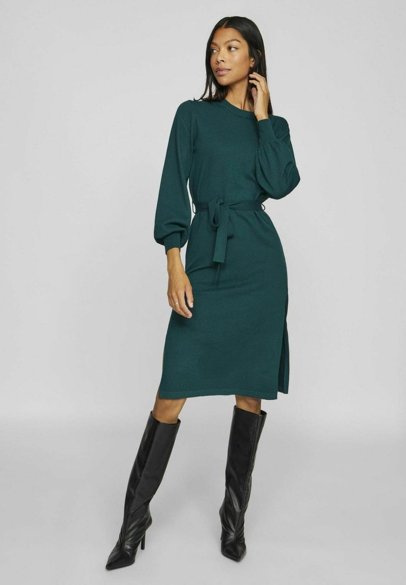 Rochie tricotată verde cu mâneci puff lungi, talie legată, fante laterale și lungime midi, asortată cu cizme negre cu vârful ascuțit până la genunchi.