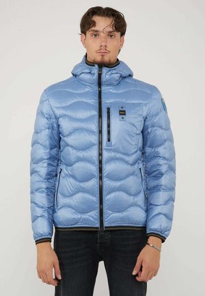 Uomo che indossa una giacca trapuntata azzurro chiaro con dettagli con zip, in piedi davanti a uno sfondo chiaro uniforme, rivolto in avanti.