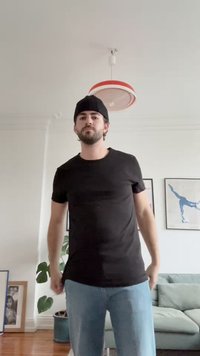 Crna majica kratkih rukava u kombinaciji s svijetloplavim trapericama. Na glavi se nosi beanie. U pozadini se nalaze sobne biljke i okvirne umjetničke slike.