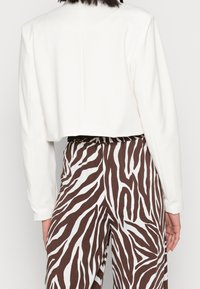 Nainen, jolla on yllään lyhyt valkoinen pitkähiha, ja korkeavyötäröiset ruskeat ja valkoiset seeprakuosiset housut, nähtynä takaa.