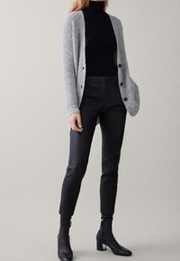 Frau in einer grauen Strickjacke, einem schwarzen Rollkragenpullover, eng anliegenden schwarzen Hosen und schwarzen Absatzstiefeletten, die vor einem schlichten Hintergrund steht.