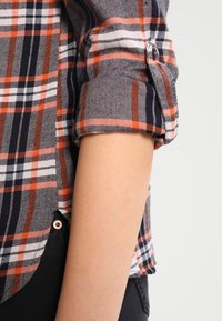 Camicia a quadri con colori blu navy, arancione e bianco; maniche arrotolate; orlo rotondo; e un singolo bottone sul polsino.