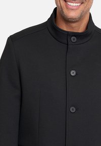 Abrigo negro texturizado con cuello alzado, tapeta frontal con dos botones y diseño ajustado. Sin patrones ni acentos visibles.