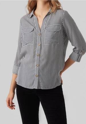 Femme portant une chemise boutonnée à rayures verticales noires et blanches avec deux poches poitrine à rayures horizontales et manches retroussées.
