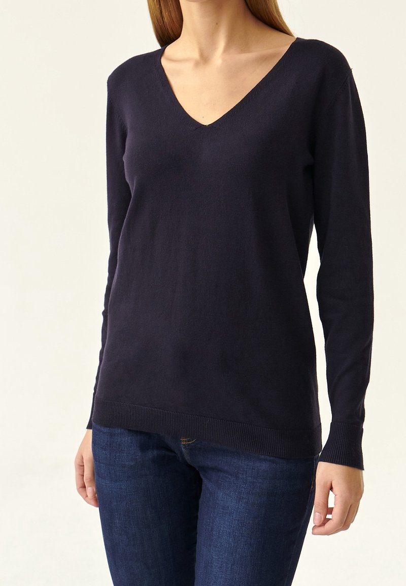 Femme portant un pull à col en V uni bleu marine foncé à manches longues, associé à un jean bleu foncé, debout devant un fond clair.