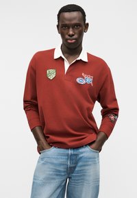 Κόκκινο μακρυμάνικο πόλο μπλουζάκι με λευκό γιακά, με τρεις κεντημένες επιδιορθώσεις και χαλαρή εφαρμογή, συνδυασμένο με ανοιχτό μπλε τζιν παντελόνι.