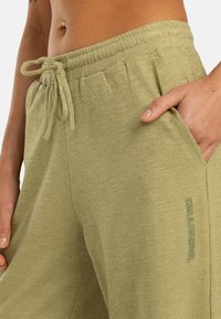 Joggers vert clair texturé avec une taille élastique, un cordon de serrage, et des poches latérales ; présente un logo imprimé sur le devant.