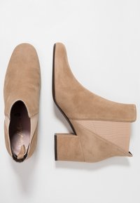 Beiga mockasklackar som når ankeln med en klack, har elastiska sidopaneler och en slät yta, vilket ger en strömlinjeformad och modern design.