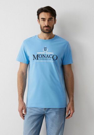 Mężczyzna w jasnoniebieskim T-shircie z napisem „MONACO Diverse Sport Club True Brand since 1993” i jasnoniebieskich dżinsach stoi przy białej ścianie.