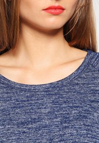 Niebieski, teksturowany top z szerokim, prążkowanym dekoltem, z mieszanym wzorem i gładkim wykończeniem. Zbliżenie na obszar dekoltu.