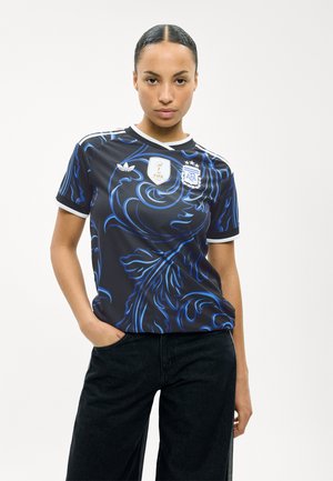 Kobieta z zaczesanymi do tyłu włosami, ubrana w czarno-niebieską koszulkę piłkarską Argentyny z logo FIFA i AFA, pozująca z jedną ręką w kieszeni.