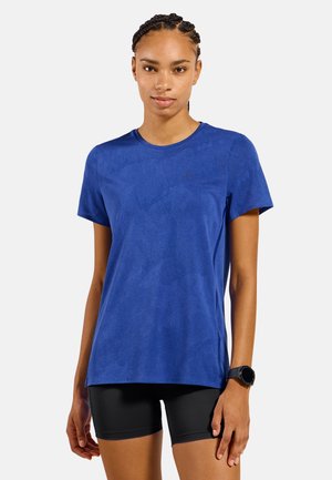 Jonge vrouw staand, draagt een blauw T-shirt met korte mouwen, zwarte fietsbroek en een zwart smartwatch aan haar linkerpols.