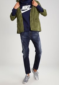 Olive green jacket over a navy hoodie with white logo, dark blue jeans, and silver sneakers. Features pockets and a relaxed fit.