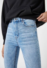 Jean en denim bleu clair avec une coupe slim, présentant un effet usé subtil, un patch déchiré et un style classique à cinq poches.