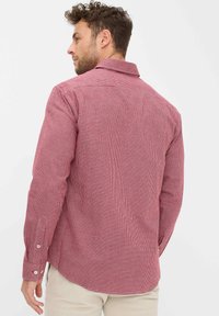 Uomo che indossa una camicia a maniche lunghe a scacchi rossi e bianchi, di spalle, abbinata a pantaloni beige chiaro, su uno sfondo neutro.