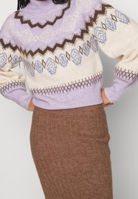 Kvinna som bär en lila, krämfärgad och brun mönstrad tröja instoppad i en hög midja ribbat brun stickad kjol.