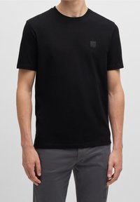 Svart bomullströja med rund hals, korta ärmar och en liten rektangulär logotyp på vänster bröst. Slät textur.