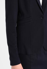 Närbild av en person som bär en mörk marinblå kavaj med en svart knapp och två horisontella fickor på sidan.