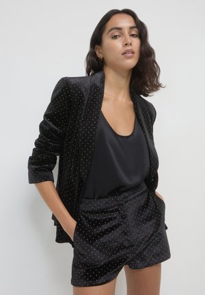 Woman with shoulder-length dark hair wearing a black silk top and black velvet blazer and shorts with small gold dots, hands in pockets.