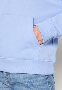 Hand i fickan på en ljusblå sweatshirt, bärs med urtvättade blå denimjeans, visar ribbat manschett och sömnadsdetaljer.