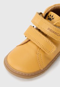 Pantofle galbene din piele cu două curele Velcro, vârf rotunjit și talpă din cauciuc brun. Prezintă un logo cu amprenta unei labe întunecate și detalii de cusătură vizibile.