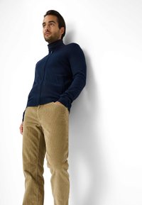 Maglione zip-up blu navy con maniche lunghe, polsi a coste e due tasche frontali, abbinato a pantaloni di velluto a coste marrone chiaro con tessuto testurizzato.