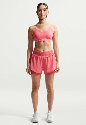 Kvinde står oprejst iført pink Nike sports-bh og løbeshorts med hvide sneakers mod en ensfarvet baggrund.