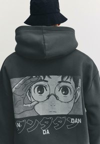 Sötétszürke kapucnis pulóver, nagy nyomattal egy szemüveges karakterről, fekete-fehér körvonalakkal, alatta japán szöveges díszítéssel.